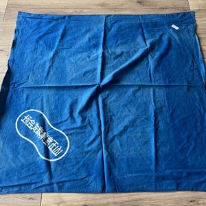 Op de afbeelding: Een vierkante blauwe stoffen doek met een witte ovaalvormige afbeelding met Japanse tekens in de linkerbenedenhoek. Het textiel heeft een licht gekreukte textuur en een diepe indigokleur over het gehele oppervlak.