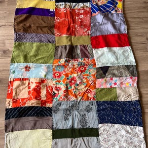 May include: A rectangular patchwork quilt constructed from various fabric squares and rectangles. The textile features a diverse array of colours including red, orange, blue, green, brown, and yellow, with patterns ranging from floral prints to geometric designs.