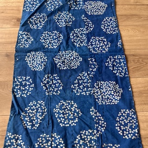 Op de afbeelding: Een rechthoekig blauw textiel met een herhalend wit bloemenpatroon van bloemtrossen. De stof vertoont over het gehele oppervlak zichtbare verticale rijen donkerblauwe rijgsteken, wat zorgt voor een doorgestikt uiterlijk.