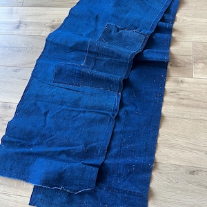 Op de afbeelding: Een rechthoekig stuk diep indigoblauwe stof met een grove geweven textuur. Het textiel heeft zichtbare stiksels langs de randen en een centraal opgestikt gedeelte, met variaties in kleurverzadiging en een rauwe, onafgewerkte zoom.
