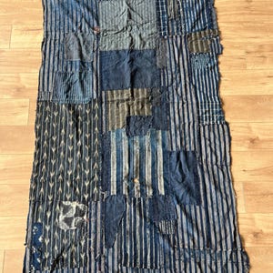 May include: A rectangular textile composed of various indigo-dyed fabric patches stitched together. The piece features a mix of vertical stripes, arrow-like patterns, and solid blue sections with visible frayed edges and worn, distressed textures.