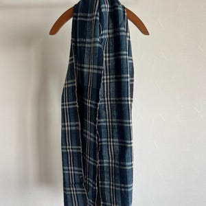 May include: A long rectangular scarf featuring a dark blue, teal, and white plaid pattern. The fabric has a visible woven texture with frayed edges along the sides, draped over a wooden hanger on a metal stand.