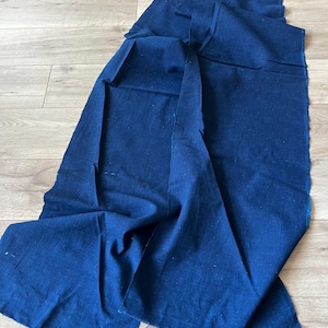Può includere: Un lungo pezzo rettangolare di tessuto blu scuro con una trama visibile. Il materiale è drappeggiato in pieghe morbide, mostrando bordi grezzi e non rifiniti lungo i lati. Il colore indaco profondo è uniforme su tutta la lunghezza del tessuto.