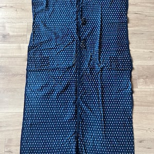 Può includere: Un pezzo di tessuto rettangolare blu scuro con un motivo ripetitivo di piccoli punti bianchi. Il tessuto presenta bordi grezzi non rifiniti e una cucitura verticale visibile al centro, con due tasche quadrate applicate sui lati.