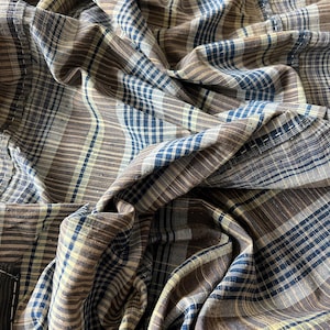 Può includere: Un tessuto con motivo a quadri caratterizzato da strisce marroni, blu e gialle. Il materiale è arricciato e piegato, creando ombre e riflessi profondi che enfatizzano la consistenza del tessuto intrecciato e le linee intersecanti del design.