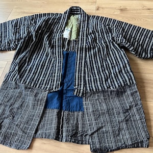 May include: A black and white striped haori jacket featuring a patchwork design with a solid blue panel on the interior and a light green fabric detail near the collar. The garment has wide sleeves and an open front, showcasing a mix of textures.