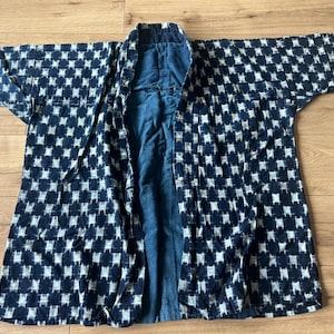 Könnte beinhalten: Eine dunkelblau-weiß gemusterte Jacke mit wiederkehrendem geometrischem Motiv. Das Kleidungsstück hat weite Ärmel, eine offene Vorderseite und ein einfarbig dunkelblaues Innenfutter. Der Stoff hat eine strukturierte Optik mit sichtbarem Gewebe.