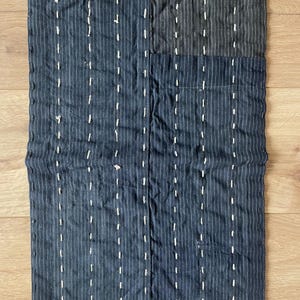May include: A rectangular textile piece featuring dark navy blue fabric with thin white pinstripes. The material is quilted with vertical rows of white running stitches, creating a textured surface. One corner displays a darker, solid navy patch.