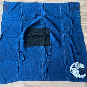 Può includere: Un pezzo di tessuto rettangolare blu con una grande toppa centrale in tessuto nero e un emblema circolare bianco nell'angolo in basso a destra. Il tessuto presenta cuciture visibili, rinforzi cuciti e una consistenza usurata.