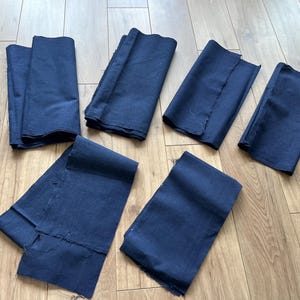 May include: A collection of folded navy blue fabric pieces, of varying sizes and shapes, are displayed on a light brown wooden floor. Some pieces have raw edges, indicating they have been cut. The fabric's deep blue hue suggests it could be used for clothing or home decor projects.