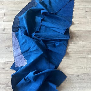 May include: A deep blue fabric panel featuring a dark, reinforced waistband and a lighter blue, rectangular patch stitched onto the side. The textile displays a visible weave and folds, with raw edges and visible stitching along the structural seams.