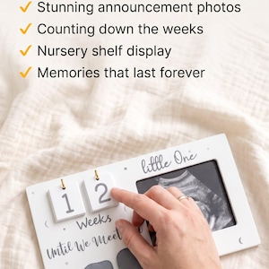 May include: A white rectangular wooden frame featuring two flip-style number blocks displaying 12, the text "little One" and "Weeks Until We Meet", and three grey elephant icons. A black and white ultrasound image is visible in the frame.