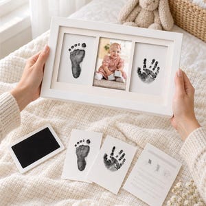 May include: A white rectangular triple-frame containing a black footprint, a photograph of a baby, and a black handprint. Below are a black ink pad, two separate cards with a footprint and handprint, and an instruction card with diagrams.