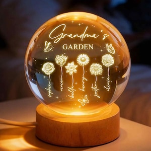 May include: A glowing glass sphere with the words "Grandma's GARDEN" and names of loved ones. The sphere features illuminated floral designs and rests on a wooden base, creating a warm, inviting ambiance.