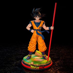 Puede incluir: Una detallada figura de Goku de Dragon Ball Z. La figura está en una pose dinámica, vistiendo un gi naranja con una faja y muñequeras azules. Sostiene un bastón rojo. La base presenta el logo de Dragon Ball.