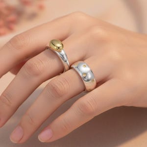 以下が含まれることがあります： 手には2つのシルバーリングが表示されています。1つのリングには、金色の楕円形の石が付いています。もう一方のリングは、小さな金の星のアクセントが付いたシルバーです。リングは、柔らかくニュートラルな背景の手の上にあります。