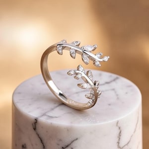 Op de afbeelding: Een zilverkleurige ring met een bladeren design. De ring heeft twee blad takken die zich om de vinger wikkelen, versierd met kleine, heldere edelstenen. De ring wordt getoond op een witte marmeren sokkel tegen een warme, neutrale achtergrond.