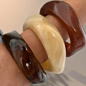 以下が含まれることがあります： 茶色とクリーム色の4つの分厚い樹脂製バングルが手首に重ねられています。 ブレスレットは光沢仕上げで、正方形で丸みを帯びた形状をしています。 色は濃い茶色からクリーミーなベージュまであります。