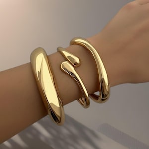 Pode incluir: Conjunto de três pulseiras douradas. As pulseiras têm um acabamento liso e polido. Uma é uma pulseira grossa e arredondada, outra é um design fino e aberto, e a terceira tem uma forma fluida única. Joias brilhantes.