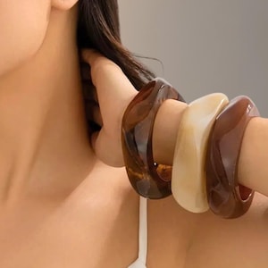 Op de afbeelding: Drie dikke, vierkante armbanden in bruin- en crèmetinten. De armbanden worden om de pols gedragen en tonen hun gladde, glanzende afwerking. Mode-accessoire.