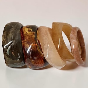 Pode incluir: Uma coleção de cinco pulseiras de resina em tons de castanho, âmbar e pêssego. As pulseiras têm um acabamento brilhante e variam em largura e forma, mostrando padrões e texturas únicos. As pulseiras estão empilhadas numa superfície branca.