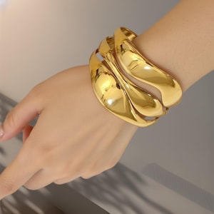 Può includere: Un set di tre braccialetti dorati, larghi e curvi. I braccialetti sono lisci e lucenti, riflettendo la luce. I gioielli sono indossati su un polso, mostrando il design e il colore.