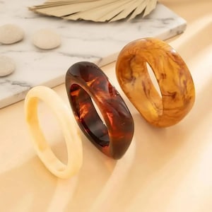Puede incluir: Tres brazaletes gruesos de resina en crema, marrón oscuro y ámbar. Los brazaletes tienen un acabado liso y brillante y una forma rectangular redondeada. El brazalete ámbar tiene un patrón veteado. Los brazaletes se exhiben sobre una superficie neutra.