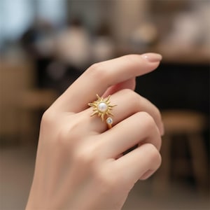 May include: A gold-colored ring featuring a sunburst design with a central pearl and a small, clear gemstone. The ring's design includes radiating rays around the pearl, and a delicate band. The ring is a piece of jewelry.