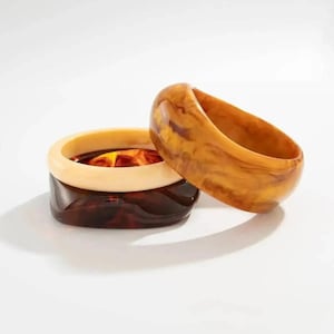 Puede incluir: Tres pulseras gruesas de resina en tonos marrón, ámbar y crema. Las pulseras tienen un efecto veteado, con un acabado liso y brillante. Las pulseras están apiladas, mostrando sus diferentes colores y texturas. Joyería.