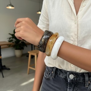 Op de afbeelding: Drie dikke armbanden in bruin, beige en wit zijn op een pols gestapeld. De armbanden hebben een gladde, gepolijste afwerking. De persoon draagt een wit overhemd met knopen en een donkere spijkerbroek.