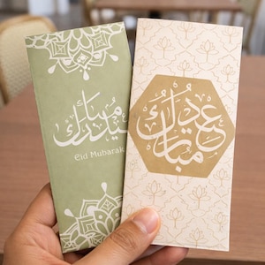 Puede incluir: Dos tarjetas rectangulares de Eid Mubarak se sostienen en la mano. Una es verde claro con diseños florales blancos y escritura árabe. La otra es blanquecina con un hexágono dorado y escritura árabe. Las tarjetas son para la festividad islámica de Eid.