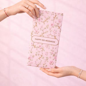 Könnte beinhalten: Eine rosa Karte mit floralem Muster und dem Text "HAPPY EID MUBARAK" wird zwischen zwei Händen gehalten. Die Karte zeigt ein sich wiederholendes Muster aus rosa Blumen und braunen Zweigen. Der Hintergrund ist ein zartes Rosa.