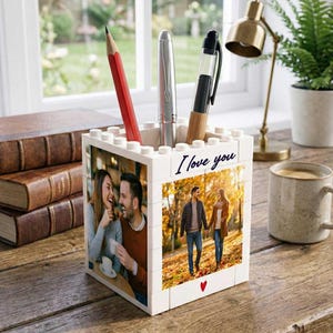 May include: A white block-style desk organizer with a photo of a couple on two sides and the words "I love you" written on the front. The organizer holds a red pencil, a silver pen, and a black pen.
