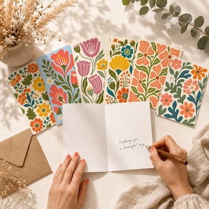 May include: A set of folded greeting cards featuring colorful floral patterns in shades of orange, pink, yellow, and blue. One card is open, displaying the handwritten text 'Wishing you a beautiful day' in black ink on the white interior page.