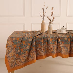 Pode incluir: Uma toalha de mesa retangular com um padrão floral em tons de verde-azulado, laranja e azul marinho. A toalha de mesa está drapeada sobre uma mesa de madeira, com vasos decorativos e um serviço de chá visíveis no topo. As bordas são laranja.