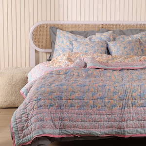 Può includere: Un letto è fatto con una trapunta blu a fantasia floreale e cuscini abbinati. La trapunta ha un bordo rosa e un design a righe. La testiera è bianca con un design in canna intrecciata. Un pouf beige è a sinistra.