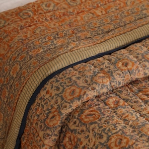 May include: Quilted bedspread with a floral pattern in shades of orange, blue, and beige. The bedding features a striped border and a dark blue trim, creating a cosy and inviting aesthetic.