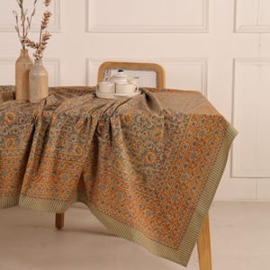 May include: A beige tablecloth with a repeating floral pattern in shades of orange and blue. The tablecloth is draped over a wooden table, with a tea set and decorative vases visible in the background. The tablecloth has a striped border.