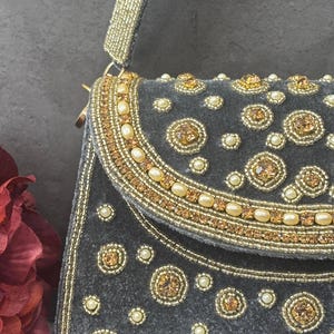 Pode incluir: Uma bolsa de mão de veludo cinza com uma alça e aba com contas. A aba e o corpo são adornados com contas douradas, pérolas falsas e strass âmbar. A bolsa tem um fecho dourado.