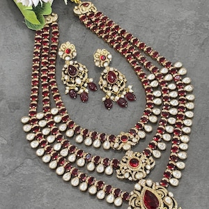 Puede incluir: Un conjunto de joyería de tres piezas con collar, pendientes y colgante a juego. El conjunto está adornado con piedras preciosas rojas y blancas engastadas en metal dorado. El collar tiene múltiples hilos y un colgante grande.