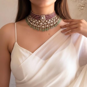 Puede incluir: Primer plano de un collar gargantilla dorado con un diseño central cuadrado y pequeñas piedras claras redondas. El collar tiene una banda de piedras rojo oscuro y una franja de piedras verdes en forma de lágrima.