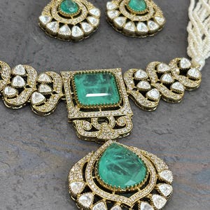May include: A gold-toned jewelry set featuring emerald-green gemstones and clear, sparkling accents. The set includes a necklace with a large square pendant, a teardrop pendant, and matching earrings. The gemstones are set in ornate, detailed frames.