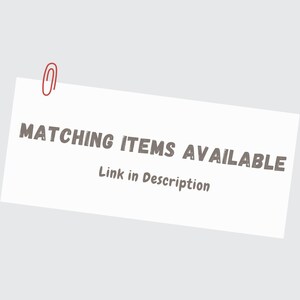 May include: A white rectangular card with the text "MATCHING ITEMS AVAILABLE" in a dark gray font. Below the text is the phrase "Link in Description". A red paper clip is attached to the top left corner of the card. The background is a light gray color.