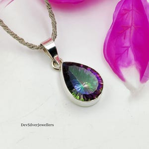 Puede incluir: Un collar con colgante de plata con una piedra preciosa iridiscente en forma de pera. La piedra preciosa muestra un espectro de colores, incluyendo verde, azul y morado. El collar está sobre un fondo blanco con acentos florales rosas. El texto "DevSilverJewellers" es visible.