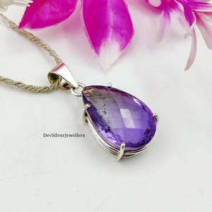 Puede incluir: Colgante de plata con una amatista facetada en forma de lágrima. La piedra púrpura tiene un efecto degradado, pasando de oscuro a claro. El colgante cuelga de una cadena de plata. La imagen está sobre un fondo blanco con flores rosas.
