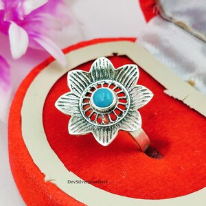 Könnte beinhalten: Ein silberner Ring mit einem Blumenmuster. Der Ring hat einen türkisblauen Stein in der Mitte, umgeben von einem detaillierten Blumenmuster. Der Ring wird in einer roten Schmuckschatulle präsentiert.