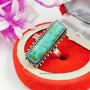 Könnte beinhalten: Ein silberner Ring mit einem rechteckigen Türkis-Edelstein. Der Türkisstein hat eine leuchtende blaugrüne Farbe und ist in einen silbernen Rahmen mit einem Perlenrand gefasst. Der Ring wird in einer roten Schmuckschatulle präsentiert. Der Text "DevSilverJewellers" ist sichtbar.