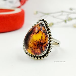Puede incluir: Un anillo de ámbar en forma de lágrima engastado en una banda de plata con un borde de cuentas. El ámbar presenta tonos naranjas y marrones profundos con inclusiones internas visibles. El texto DevSilverJewellers aparece en la esquina inferior.