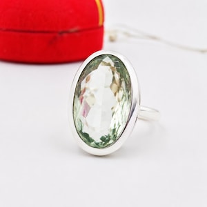 Puede incluir: Un anillo de plata con una gran piedra preciosa de color verde claro, forma ovalada y tallada en facetas, engastada en un bisel de metal liso. La piedra muestra un tono verde pálido y claro con múltiples facetas geométricas.