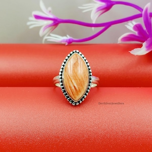 Könnte beinhalten: Ein silberner Ring mit einem ovalen, orange-braunen Edelstein. Der Stein ist in einem perlenbesetzten Rahmen gefasst. Der Ring liegt auf einer roten Oberfläche, mit lila und weißen Blumen im Hintergrund. Der Ring ist ein Schmuckstück.